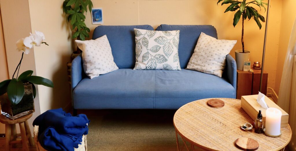 A blue sofa with patterned cushions in a warm therapy room, with plants and a round wooden table