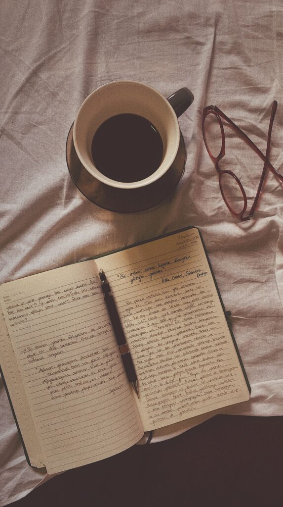Open journal with handwritten notes and a pen, alongside a cup of coffee and reading glasses, set up for a journaling self-care practice