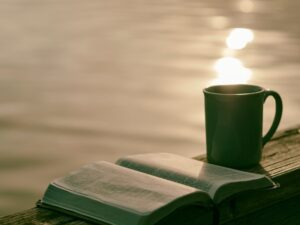 A cup of tea on a wooden ledge beside an open book, backlit by golden late-afternoon sun reflecting off rippling water.